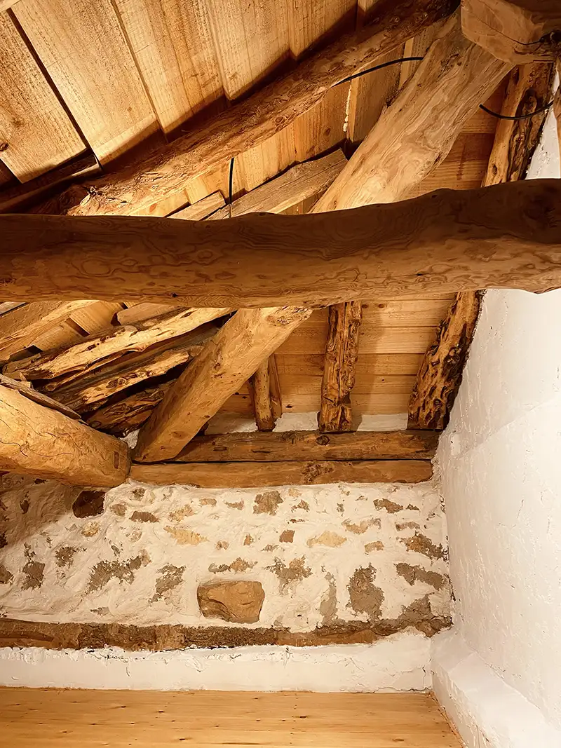 El techo de madera y las paredes de piedra blanca restauradas reflejan la esencia tradicional de la vivienda. Techo de madera restaurado en vivienda tradicional, con vigas de madera originales y paredes de piedra blanca.